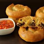 Golden-brown meat-filled pastries are arranged on a dark surface. One pastry is torn open, showing a savory filling inside. A small white bowl of vibrant red salsa with a spoon sits beside the pastries, suggesting a delicious pairing.