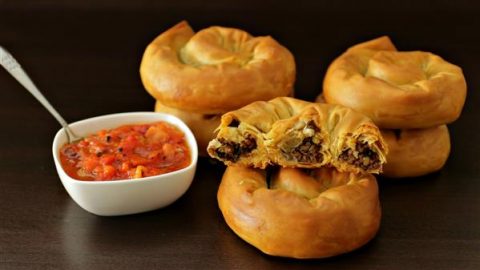 Golden-brown meat-filled pastries are arranged on a dark surface. One pastry is torn open, showing a savory filling inside. A small white bowl of vibrant red salsa with a spoon sits beside the pastries, suggesting a delicious pairing.