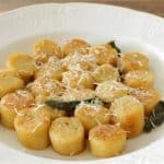 A white plate displays perfectly browned, round gnocchi pieces arranged neatly in a circular pattern. The gnocchi is garnished with shredded cheese and fresh sage leaves, providing a simple yet elegant presentation on a wooden table background.