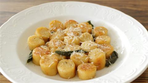 A white plate displays perfectly browned, round gnocchi pieces arranged neatly in a circular pattern. The gnocchi is garnished with shredded cheese and fresh sage leaves, providing a simple yet elegant presentation on a wooden table background.