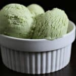 A white ribbed bowl filled with three scoops of green ice cream sits against a dark background. The ice cream appears creamy and smooth.