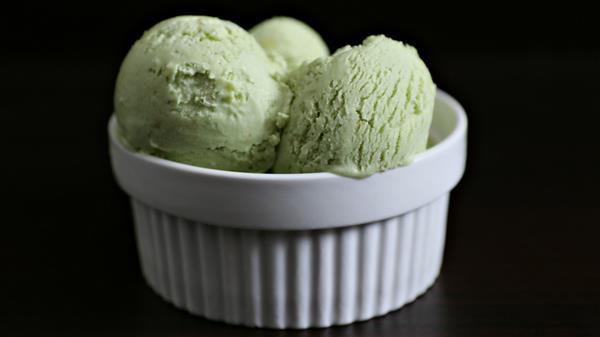 A white ribbed bowl filled with three scoops of green ice cream sits against a dark background. The ice cream appears creamy and smooth.