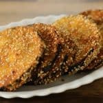 Crispy golden-brown eggplant slices coated in breadcrumbs and sesame seeds are arranged neatly on a white scalloped plate, with a wooden surface in the background.