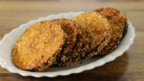 Crispy golden-brown eggplant slices coated in breadcrumbs and sesame seeds are arranged neatly on a white scalloped plate, with a wooden surface in the background.