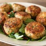 A white plate with several golden-brown salmon patties arranged on a bed of fresh greens, placed on a wooden table. The patties are garnished with herbs and have a crispy, textured exterior.