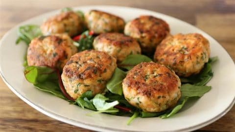 A white plate with several golden-brown salmon patties arranged on a bed of fresh greens, placed on a wooden table. The patties are garnished with herbs and have a crispy, textured exterior.