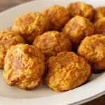 A white oval plate holds several round, golden-brown baked falafel balls. The falafel balls have a slightly crispy exterior with a textured surface, indicating a mix of ingredients and spices. The plate is placed on a wooden table.