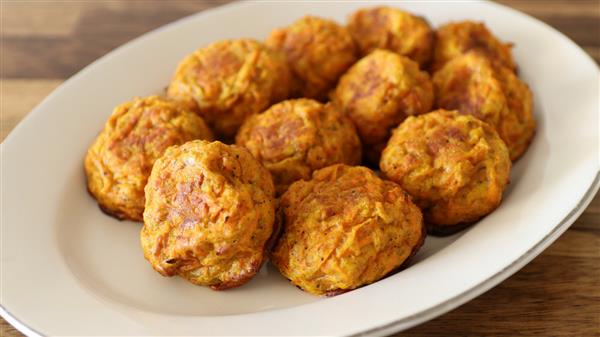 A white oval plate holds several round, golden-brown baked falafel balls. The falafel balls have a slightly crispy exterior with a textured surface, indicating a mix of ingredients and spices. The plate is placed on a wooden table.