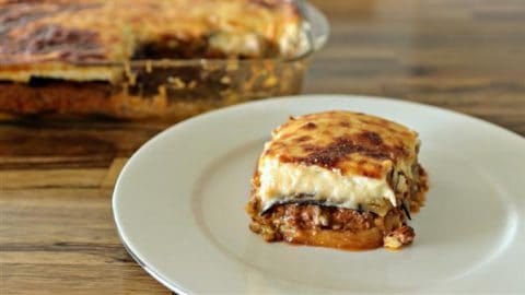 A slice of moussaka sits on a white plate. The dish features layers of browned cheese, creamy béchamel sauce, meat, and vegetables. In the background, a larger portion of the moussaka rests in a glass baking dish, with a piece missing.