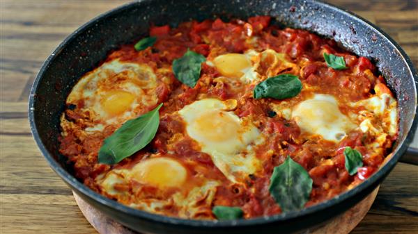 A skillet of shakshuka, a Middle Eastern dish featuring poached eggs in a rich tomato sauce with red peppers and onions, garnished with fresh basil leaves.