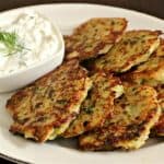 A plate of golden-brown zucchini fritters is served alongside a small dish of creamy white dip garnished with fresh dill. The fritters are arranged neatly on a white platter, and the dip sits in a square bowl, complementing the crispy appetizer.