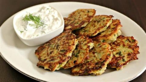 A plate of golden-brown zucchini fritters is served alongside a small dish of creamy white dip garnished with fresh dill. The fritters are arranged neatly on a white platter, and the dip sits in a square bowl, complementing the crispy appetizer.