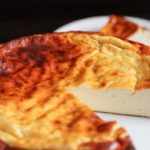 A creamy, golden-brown baked cheesecake with a slightly caramelized surface is displayed on a white plate. A single slice has been cut from the cake and placed on a smaller white plate in the background.