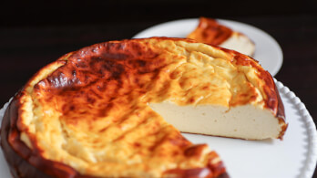 A creamy, golden-brown baked cheesecake with a slightly caramelized surface is displayed on a white plate. A single slice has been cut from the cake and placed on a smaller white plate in the background.