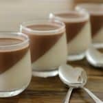 Four glass jars are filled with a dessert that has a white layer on the bottom and a brown layer on top, angled diagonally. They are arranged in a row on a wooden surface with three spoons placed beside them.