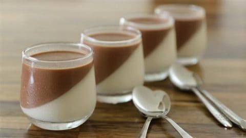 Four glass jars are filled with a dessert that has a white layer on the bottom and a brown layer on top, angled diagonally. They are arranged in a row on a wooden surface with three spoons placed beside them.