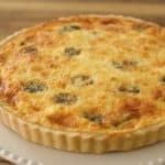 A freshly baked quiche with a golden-brown crust and a melted cheese top, speckled with green vegetable pieces, sits on a white ceramic plate with a scalloped edge. The background is a wooden surface.