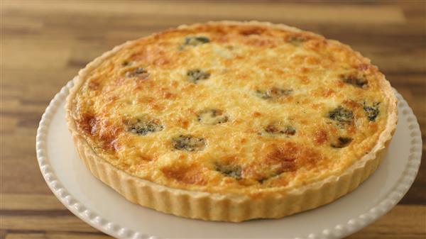 A freshly baked quiche with a golden-brown crust and a melted cheese top, speckled with green vegetable pieces, sits on a white ceramic plate with a scalloped edge. The background is a wooden surface.