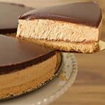 A hand uses a golden spatula to lift a slice of a chocolate-topped, cream-filled cake with a crumbly crust from a cake stand. The cake has a smooth chocolate glaze on top, a creamy middle layer, and a crunchy base.