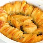 An oval baking dish filled with a beautifully arranged potato dish, featuring thin, golden-brown potato slices fanned out in a spiral pattern. The dish is garnished with sprigs of fresh herbs.