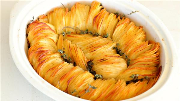 An oval baking dish filled with a beautifully arranged potato dish, featuring thin, golden-brown potato slices fanned out in a spiral pattern. The dish is garnished with sprigs of fresh herbs.