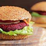 A close-up of a vegetarian beet burger with a sesame seed bun, lettuce, tomato, onion, and shredded carrots on a wooden cutting board. Another similar burger is slightly blurred in the background.