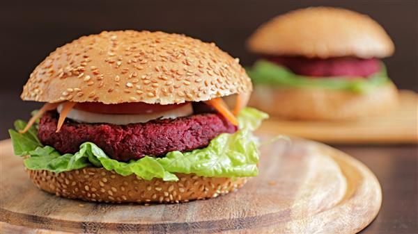 A close-up of a vegetarian beet burger with a sesame seed bun, lettuce, tomato, onion, and shredded carrots on a wooden cutting board. Another similar burger is slightly blurred in the background.