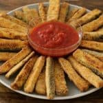 A round plate of breaded zucchini fries arranged in a radiating pattern around a small clear bowl filled with marinara sauce. The plate is on a wooden surface. The fries appear golden and crispy.