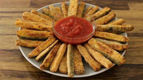 A round plate of breaded zucchini fries arranged in a radiating pattern around a small clear bowl filled with marinara sauce. The plate is on a wooden surface. The fries appear golden and crispy.