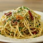 A white plate filled with spaghetti aglio e olio, topped with slices of cooked garlic, red chili flakes, fresh parsley, and grated Parmesan cheese. The pasta is lightly coated in olive oil, giving it a glossy appearance. The background is a wooden table.