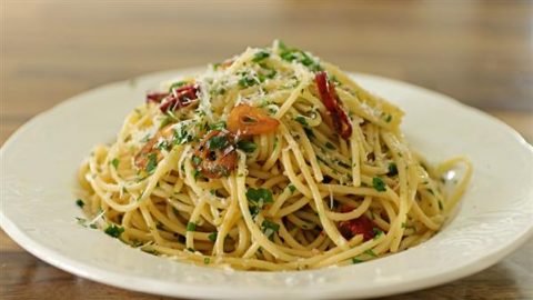 A white plate filled with spaghetti aglio e olio, topped with slices of cooked garlic, red chili flakes, fresh parsley, and grated Parmesan cheese. The pasta is lightly coated in olive oil, giving it a glossy appearance. The background is a wooden table.