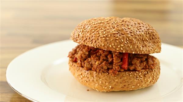 A sesame seed bun sandwich filled with seasoned ground meat is placed on a white plate. The background is a blurred wooden surface.