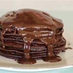 A stack of chocolate pancakes drenched in melted chocolate sauce on a white plate. The pancakes are evenly layered, and the sauce drips down the sides, creating a rich and indulgent appearance. The background is a soft, light green color.