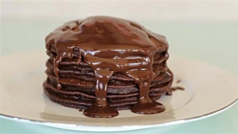 A stack of chocolate pancakes drenched in melted chocolate sauce on a white plate. The pancakes are evenly layered, and the sauce drips down the sides, creating a rich and indulgent appearance. The background is a soft, light green color.