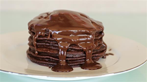 A stack of chocolate pancakes drenched in melted chocolate sauce on a white plate. The pancakes are evenly layered, and the sauce drips down the sides, creating a rich and indulgent appearance. The background is a soft, light green color.