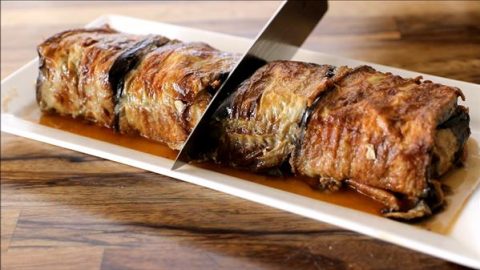 A knife slicing through a rectangular loaf of meatloaf on a white rectangular plate. The meatloaf has a caramelized crust and is covered in a rich brown sauce. The background features a wooden surface.