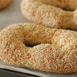 A close-up of freshly baked, oval-shaped bagels covered in sesame seeds, resting on a sheet of parchment paper. The image shows three bagels with a golden-brown crust, highlighting the texture and seed coating.