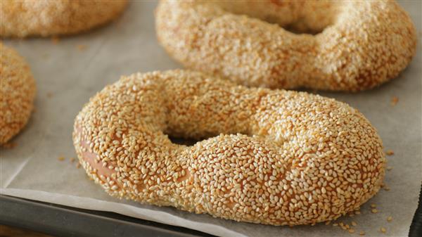 A close-up of freshly baked, oval-shaped bagels covered in sesame seeds, resting on a sheet of parchment paper. The image shows three bagels with a golden-brown crust, highlighting the texture and seed coating.