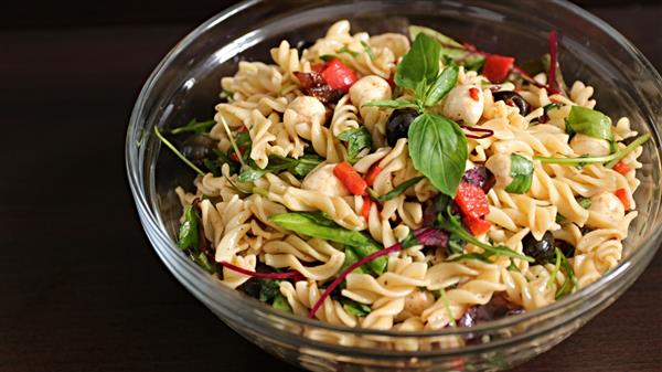 A glass bowl filled with pasta salad features rotini pasta mixed with green leaves, red bell pepper pieces, black olives, and small mozzarella balls, garnished with fresh basil leaves. The dish sits on a dark surface.