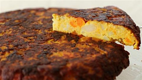 A close-up of a thick Spanish omelet, also known as a tortilla, with a slice cut and placed slightly away from the main piece. The omelet shows a golden-brown, crispy outer layer and reveals a mix of potatoes and carrots inside. The surface appears well-cooked.