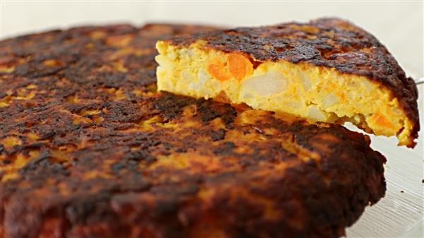 A close-up of a thick Spanish omelet, also known as a tortilla, with a slice cut and placed slightly away from the main piece. The omelet shows a golden-brown, crispy outer layer and reveals a mix of potatoes and carrots inside. The surface appears well-cooked.