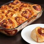 A glass baking dish filled with baked cheesy pinwheel rolls next to a white plate holding a single pinwheel roll. The rolls have a golden-brown crust with a melty cheese topping, and they are arranged in a grid inside the baking dish.