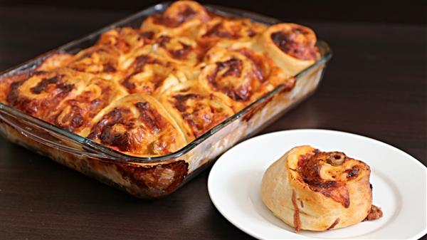 A glass baking dish filled with baked cheesy pinwheel rolls next to a white plate holding a single pinwheel roll. The rolls have a golden-brown crust with a melty cheese topping, and they are arranged in a grid inside the baking dish.