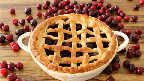 A freshly baked cherry pie with a golden, lattice crust sits on a wooden surface, surrounded by scattered cherries. The pie’s vibrant filling peeks through the lattice pattern, giving it a delicious and inviting appearance.