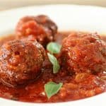 Three meatballs covered in a rich tomato sauce with a few fresh basil leaves serve as garnish. The dish is presented in a white bowl and the sauce appears thick with visible chunks of tomato.