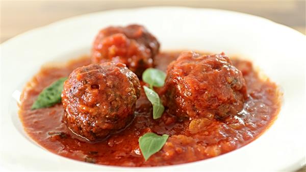 Three meatballs covered in a rich tomato sauce with a few fresh basil leaves serve as garnish. The dish is presented in a white bowl and the sauce appears thick with visible chunks of tomato.