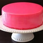 A round cake with a smooth, glossy pink frosting sits on a white pedestal cake stand. The cake is decorated with small red beads around its base. The background is a solid dark color.