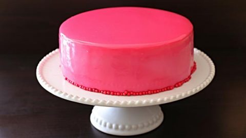 A round cake with a smooth, glossy pink frosting sits on a white pedestal cake stand. The cake is decorated with small red beads around its base. The background is a solid dark color.