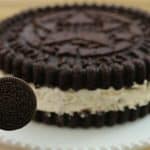A person holds an Oreo cookie in front of a large cake designed to look like an Oreo. The cake has chocolate layers on the top and bottom with white cream in between, mimicking the appearance of an Oreo cookie, placed on a white plate.