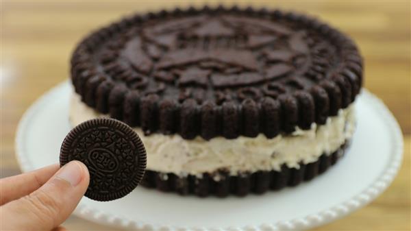 A person holds an Oreo cookie in front of a large cake designed to look like an Oreo. The cake has chocolate layers on the top and bottom with white cream in between, mimicking the appearance of an Oreo cookie, placed on a white plate.